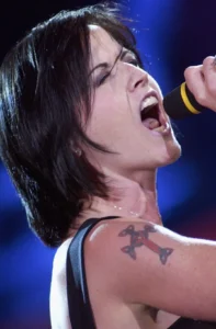Dolores O’Riordan singing on stage with her red cross shoulder tattoo clearly visible