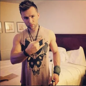 Harry Judd showing his arm tattoos while posing in a hotel room