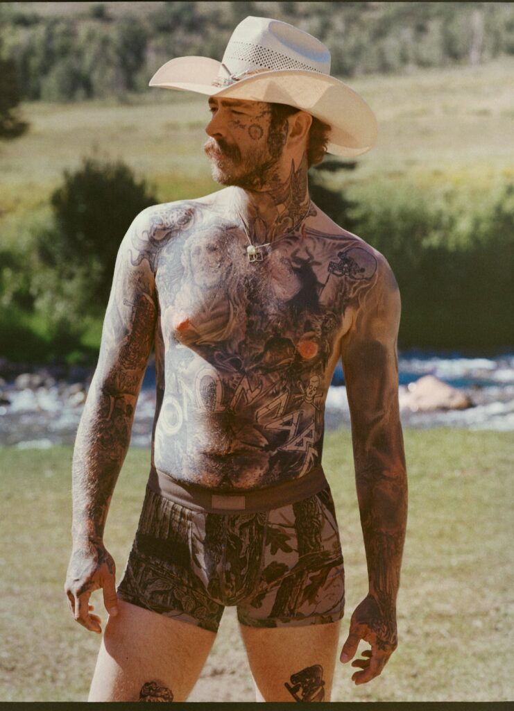 “Post Malone shirtless showing full chest, neck, and arm tattoos while wearing a cowboy hat.”